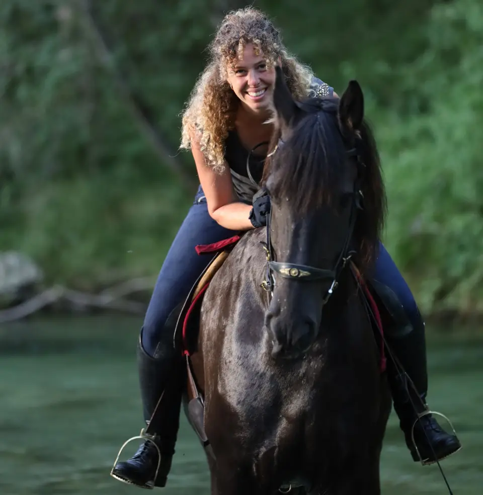 Frau mit langen Locken reitet lachend ein dunkles Pferd ohne Sattel durch einen Fluss