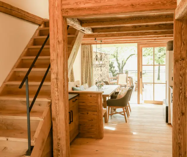 Holztreppe mit schwarzem Geländer im AlmChalet, dahinter ein Essbereich mit Holztisch vor großen Fenstern